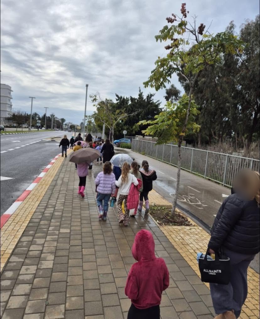 ילדים ומבוגרים הולכים במדרכה בגשם, מצולמים מאחור