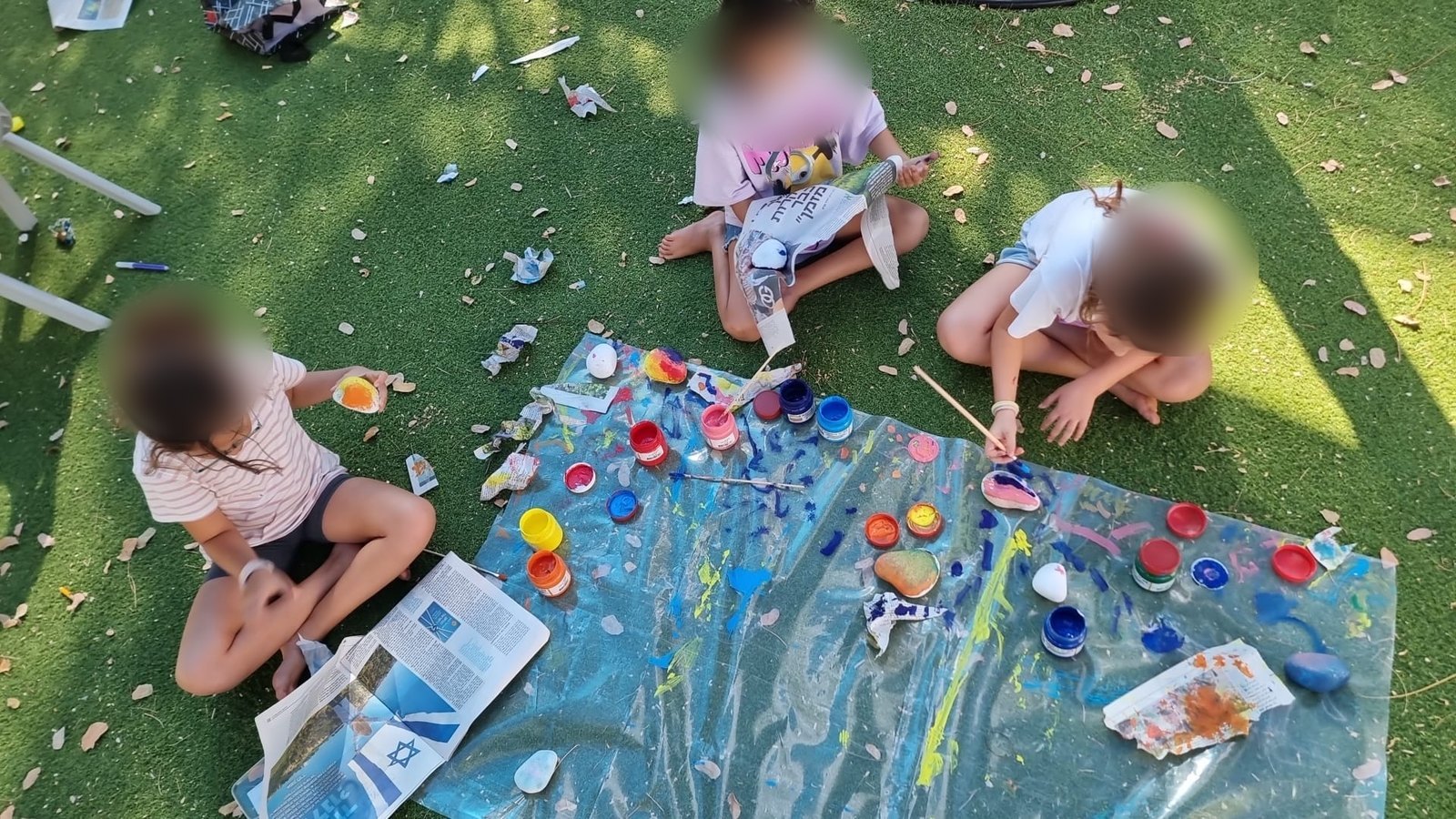 שלוש ילדות צובעות אבנים על יריעה בדשא, מלמעלה ובפנים מטושטשות